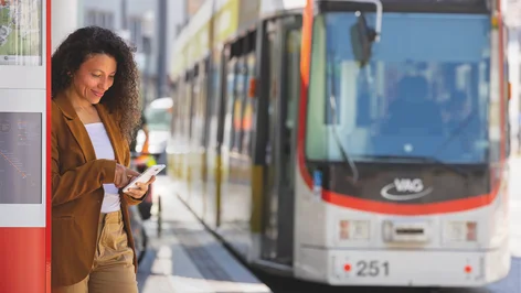 Frau an Straßenbahnhaltestelle schaut auf ihr Smartphone